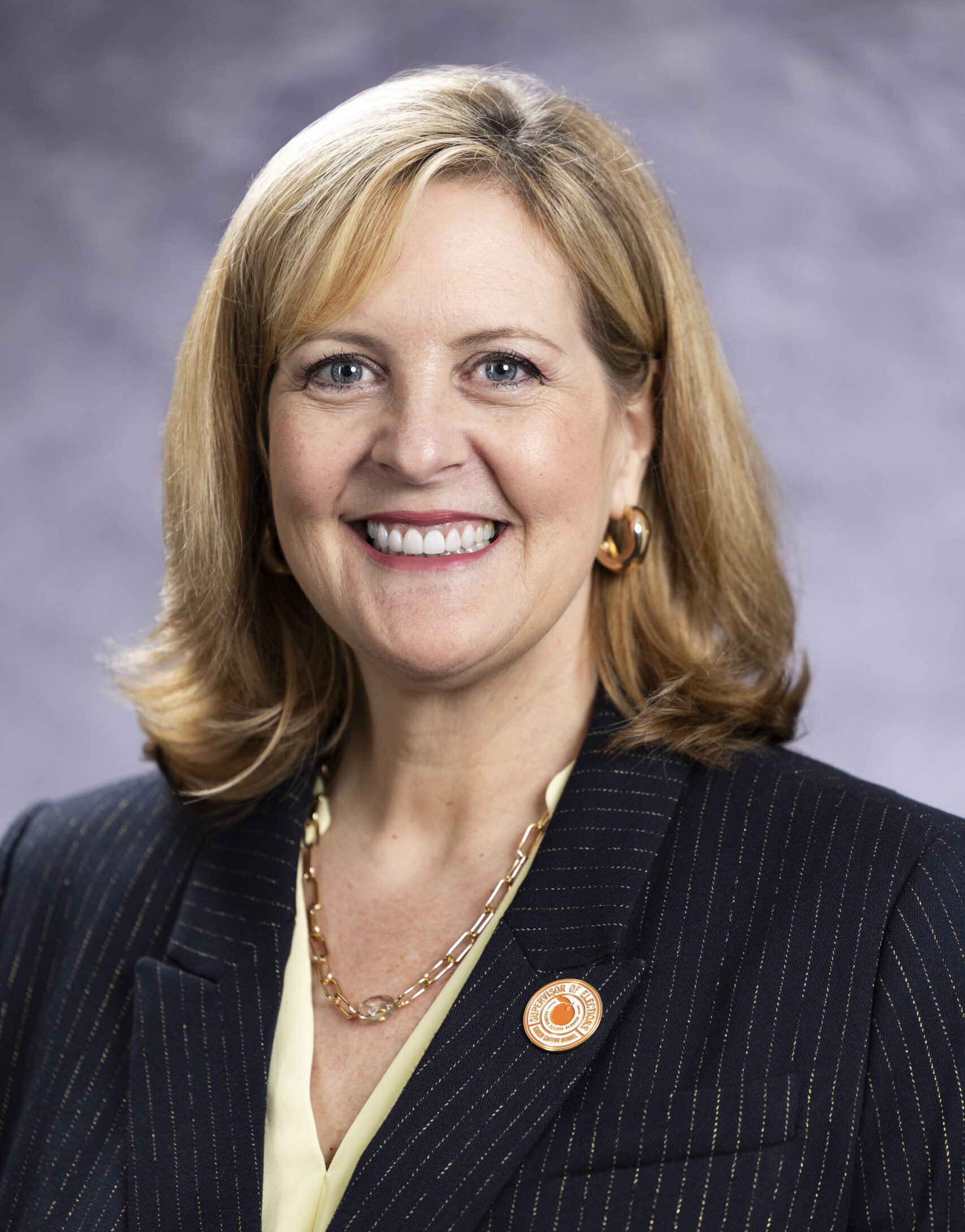 Professional headshot of a smiling woman with shoulder-length blonde hair (Karen Castor Dentel), wearing a dark pinstripe blazer, light blouse, gold jewelry, and a circular lapel pin, posed against a neutral gray background.