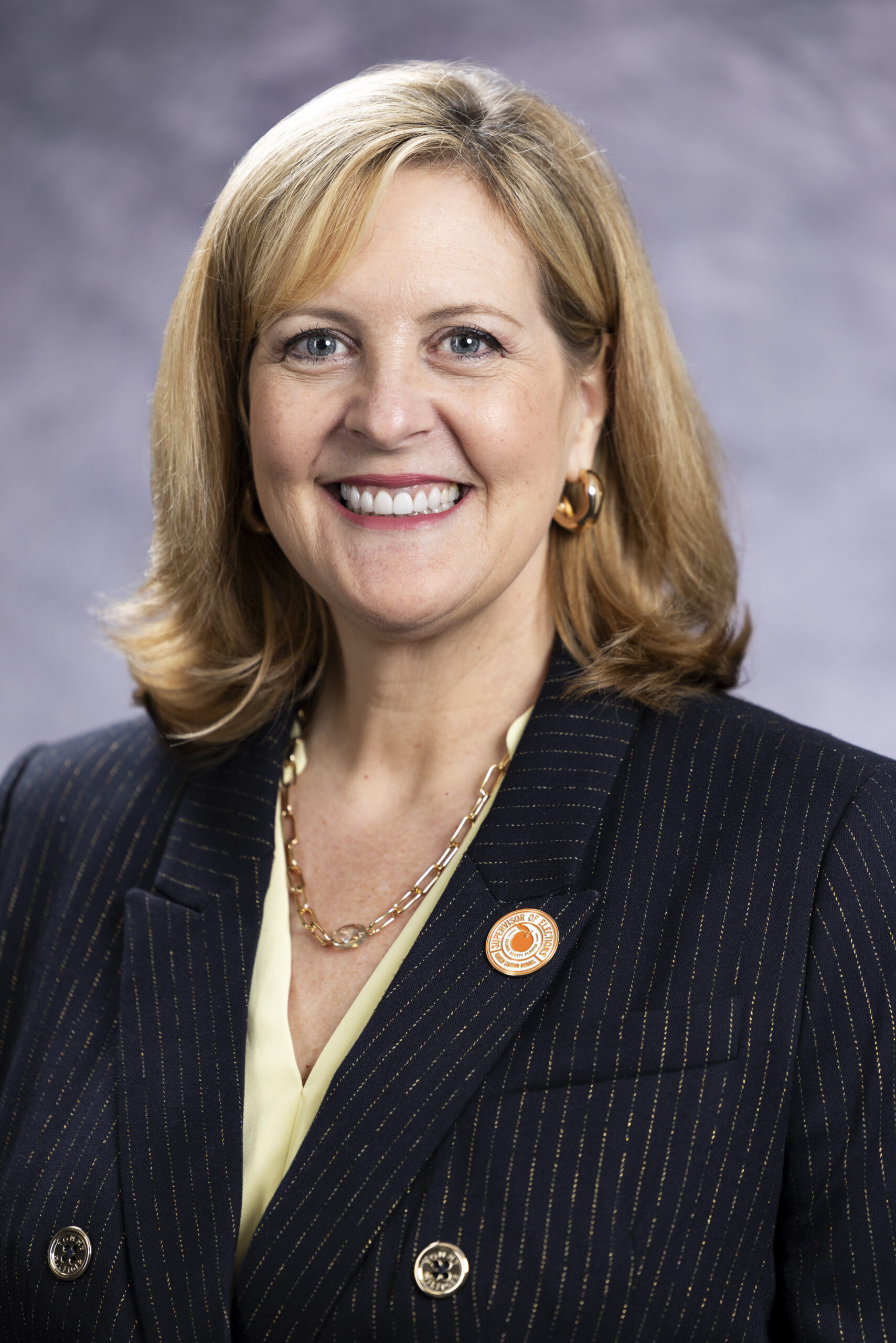 Professional headshot of a smiling woman with shoulder-length blonde hair (Karen Castor Dentel), wearing a dark pinstripe blazer, light blouse, gold jewelry, and a circular lapel pin, posed against a neutral gray background.