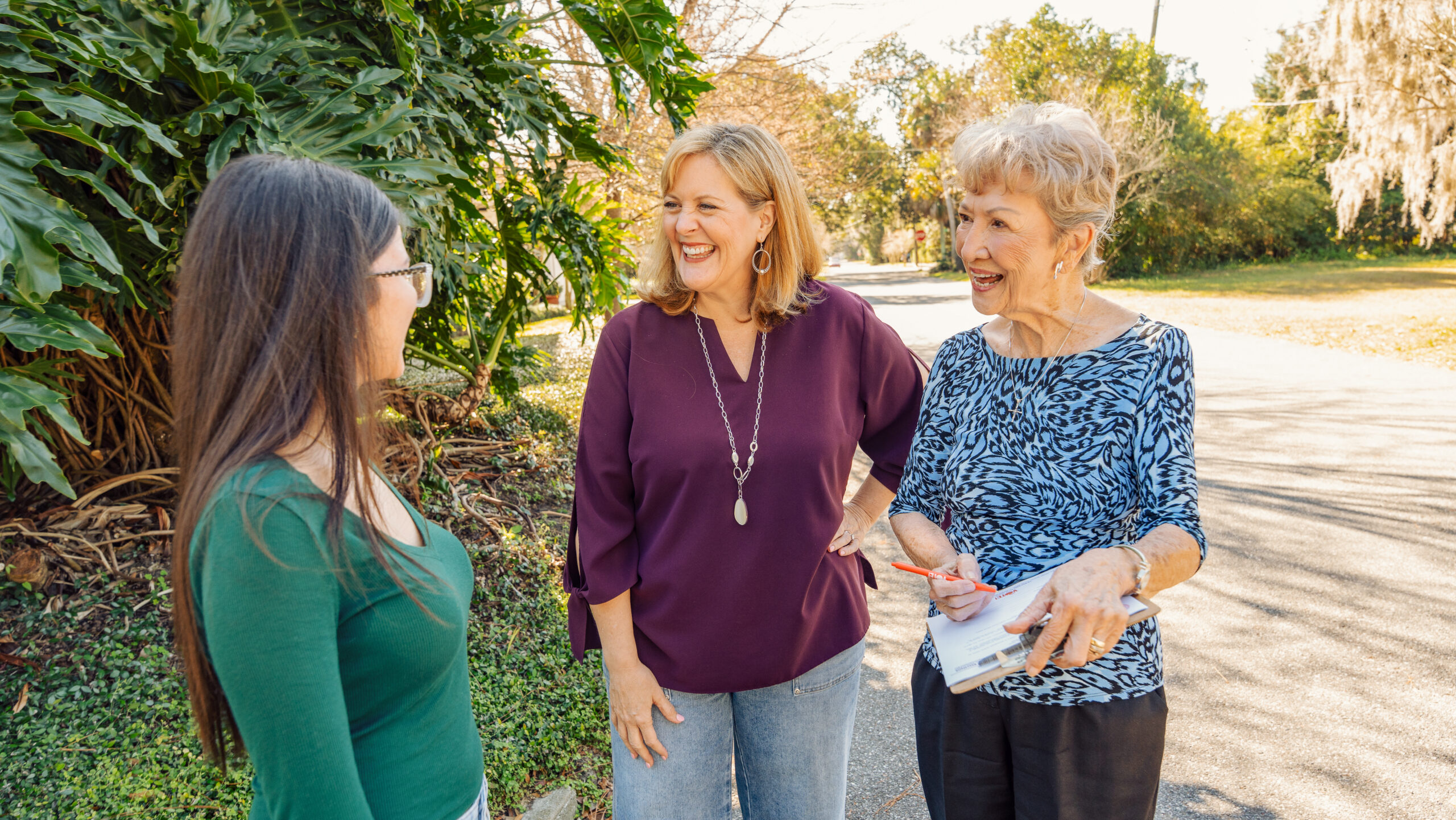 Three women stand outdoors on a sunny day, smiling and talking along a tree-lined street; one holds a clipboard and pen while speaking with the others.