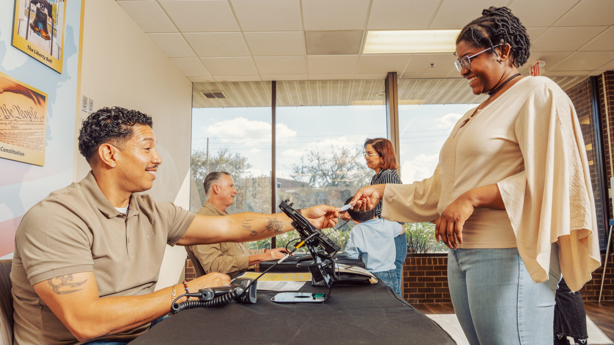 A voter hands an ID to a poll worker seated at a check-in table inside a bright polling location, while other voters and staff interact in the background near large windows.